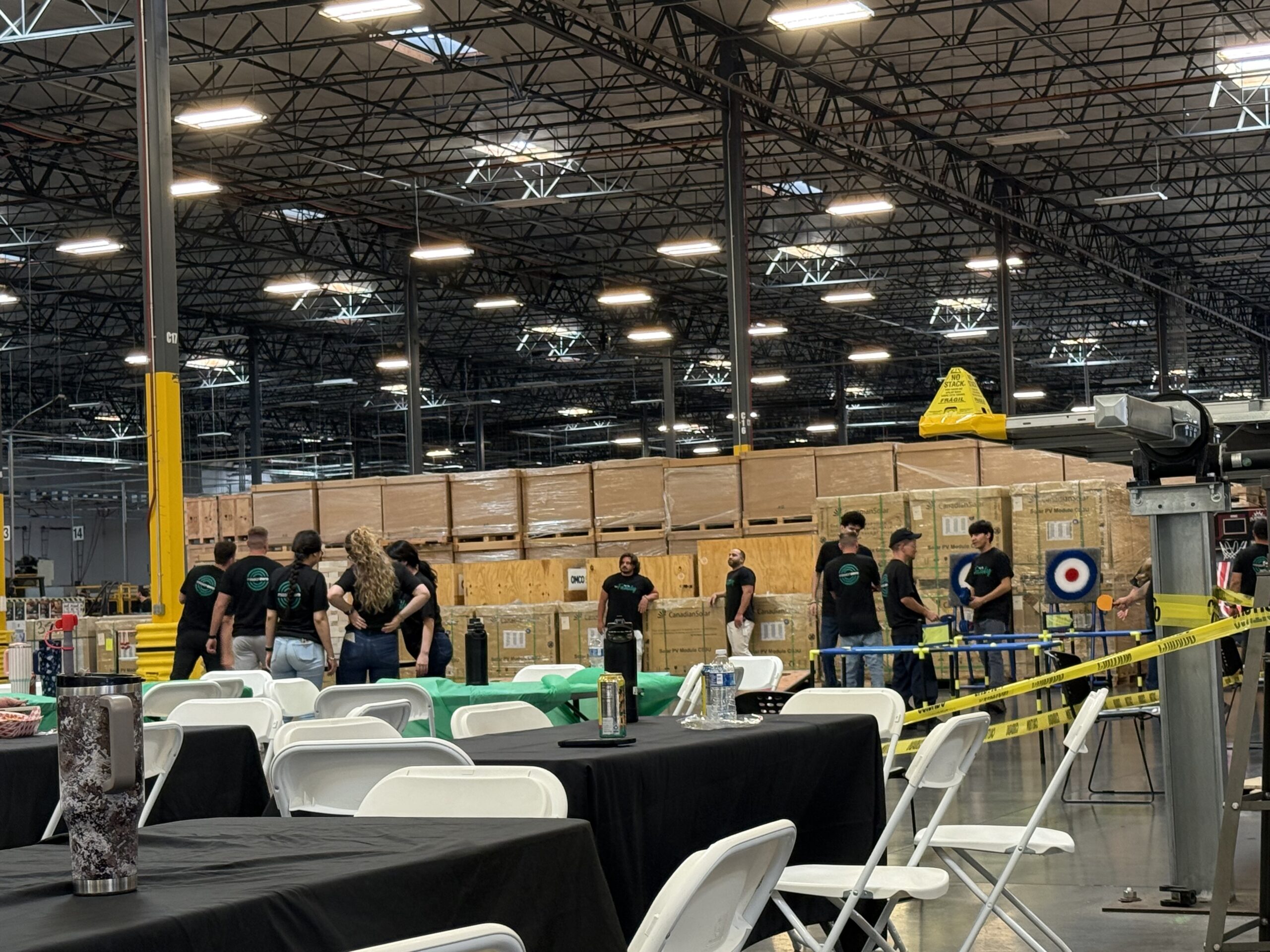 Indoor warehouse event setup with folding chairs around black-clothed tables and yellow caution tape in the foreground of a large industrial hall.